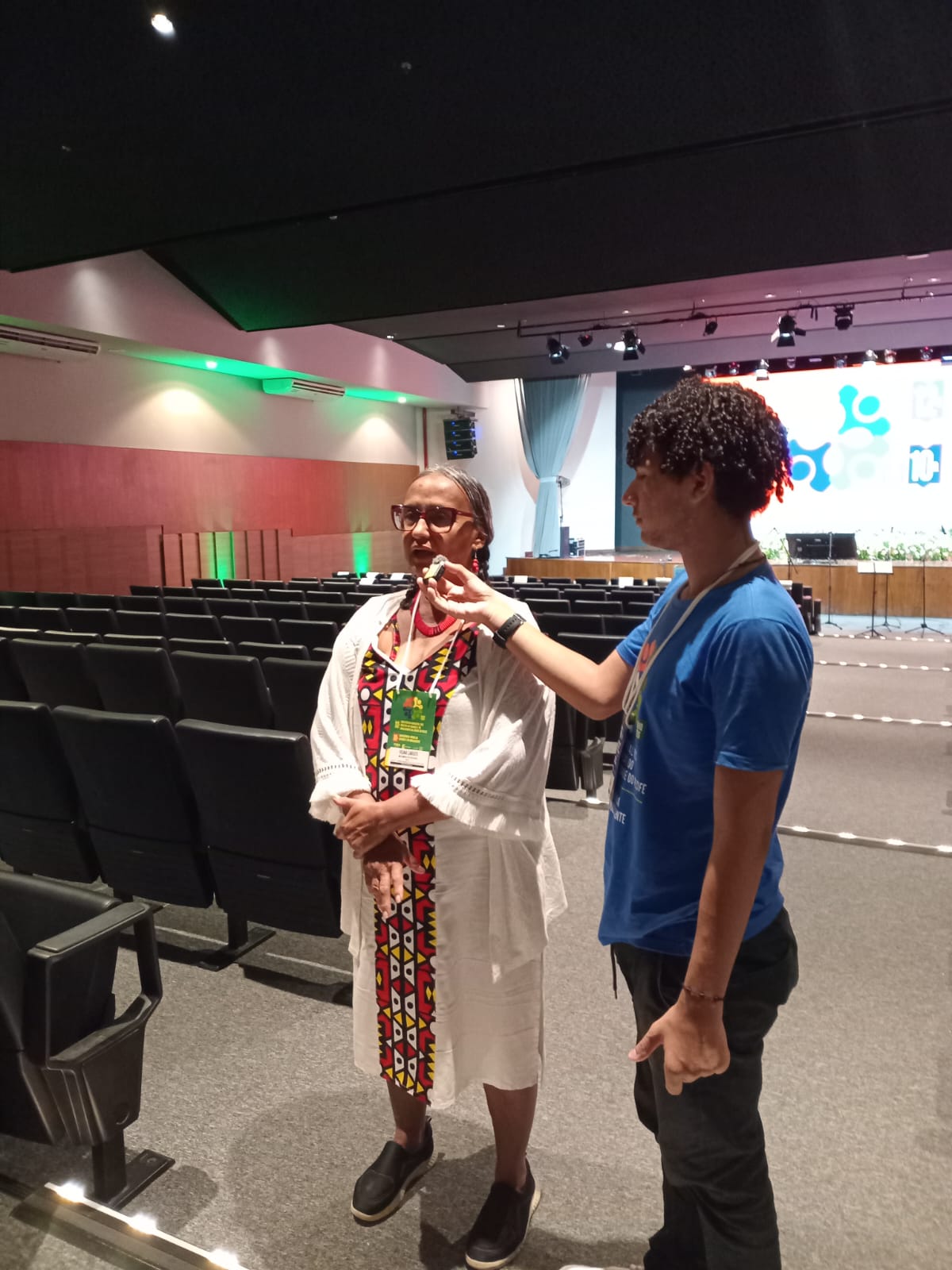 Jovem segura um pequeno microfone próximo a uma entrevistada, em um auditório.